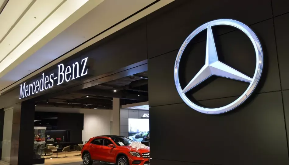 A Mercedes Benz car dealership showroom shows the partial view of a Red hatchback car. A large Mercedes logo occupies the store exterior.