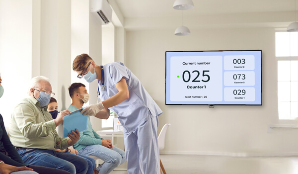 Patients sit in a hospital waiting room staring at a digital screen that displays the live queue tickets using a queue management software