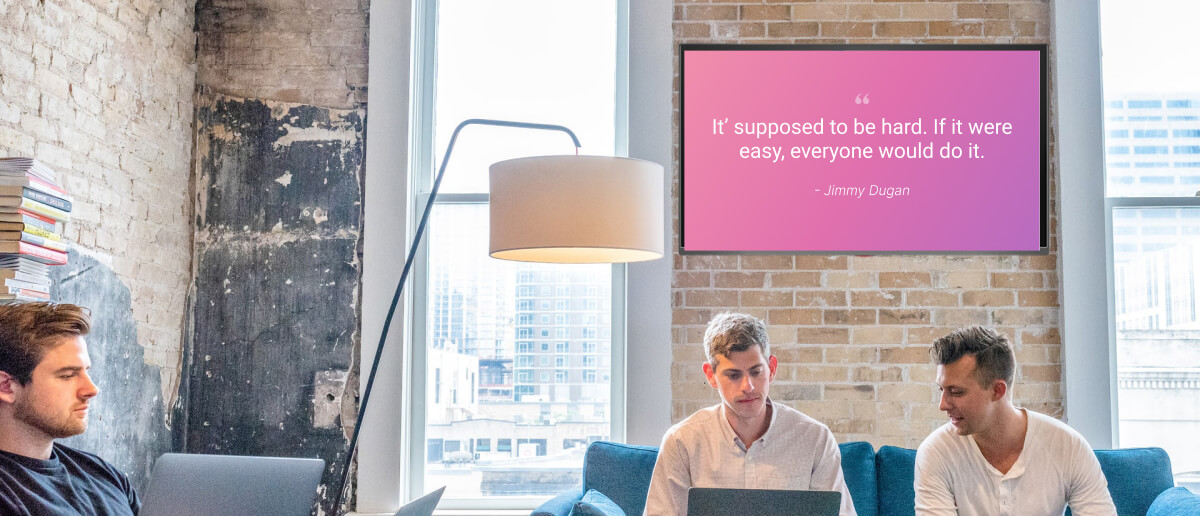 employees sit and work as a digital signage screen in the background shows company culture quotes