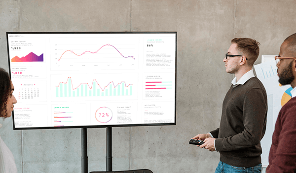A corporate team discusses a sales dashboard displayed on a digital signage inside a meeting room