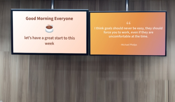 Good morning wish and motivational quote on hard work are shown on two digital signage screens of an office