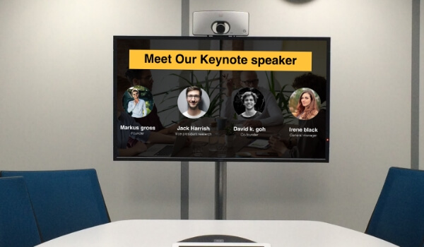 A conference room shows the names of the keynote speakers on a large digital signage display