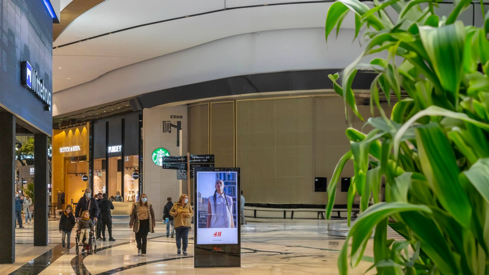 A shopping mall with people walking and a digital ad display in the center.