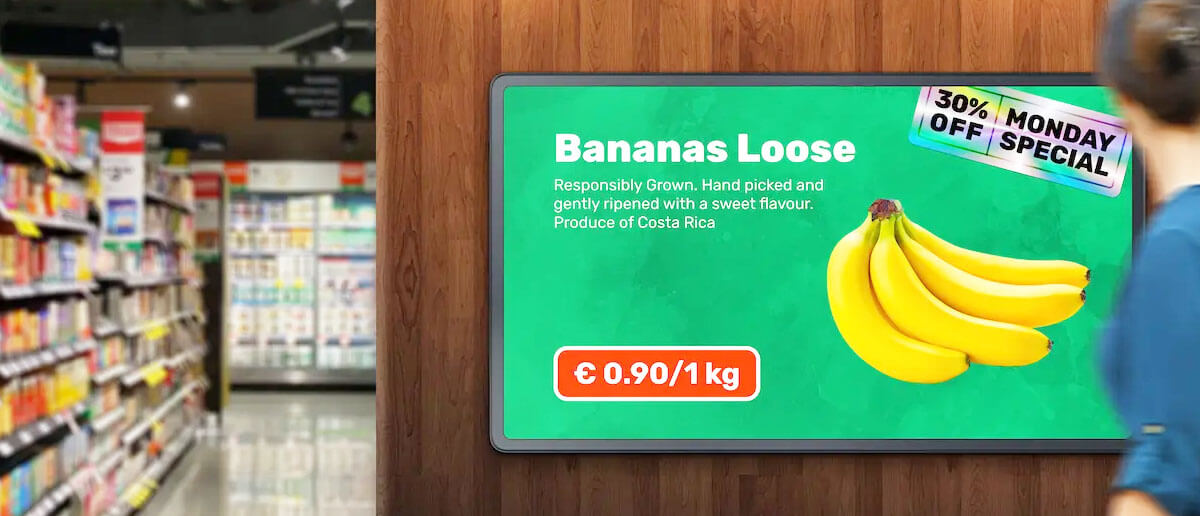 A supermarket store digital signage promotes offers on produce as a customer looks on