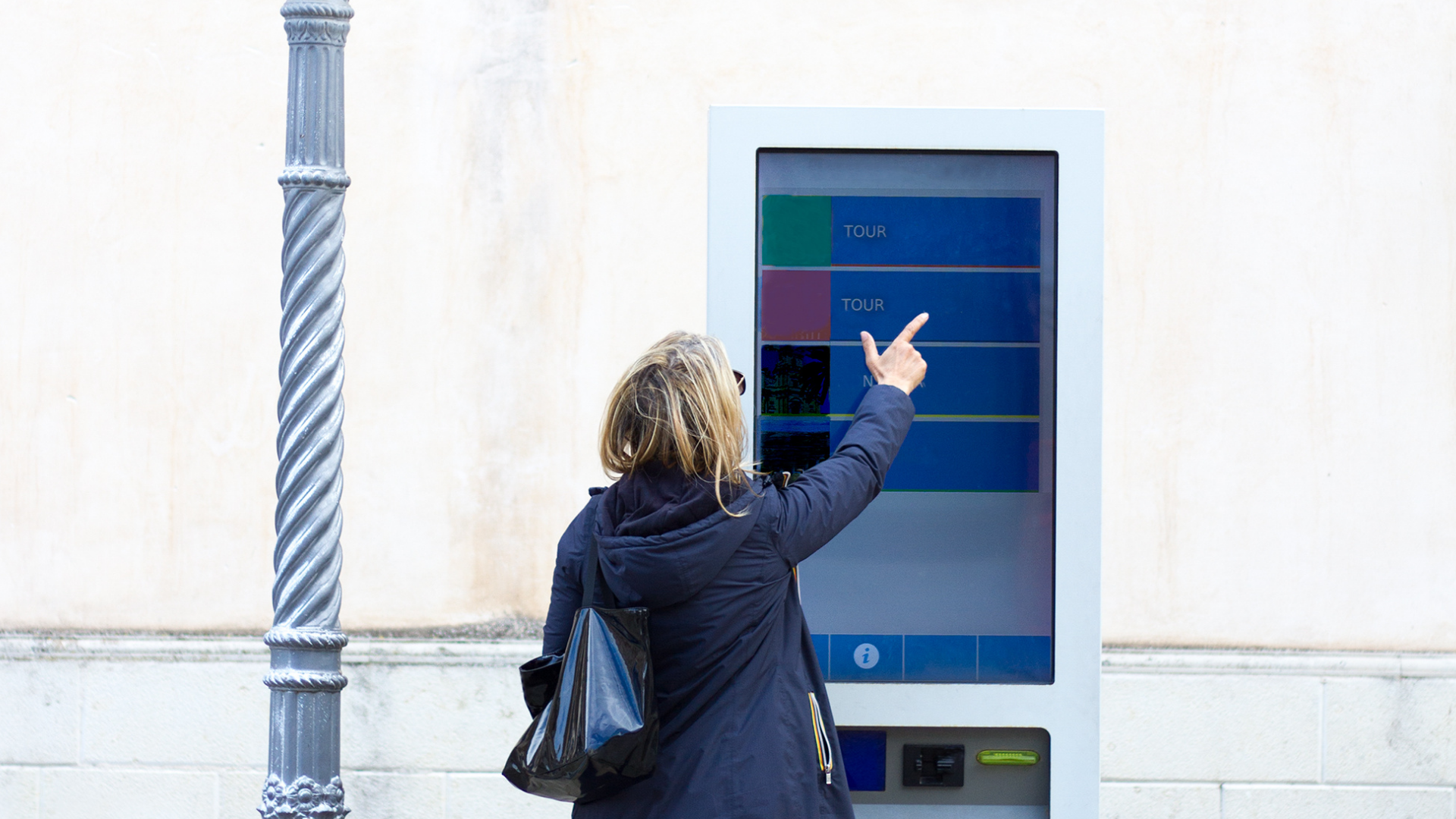 A woman dressed in a black jacket interacts with a touchscreen outdoor digital signage kiosk installed on the sidewalk of a building. The digital signage screen shows four clickable options.