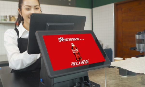A woman at a restaurant works with a point of sale billing system while the customer facing iPad shows a coca-cola ad in Chinese