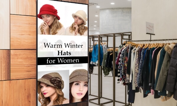 An in-store PoS digital signage display near women's winter clothing section advertises winter hats to upsell