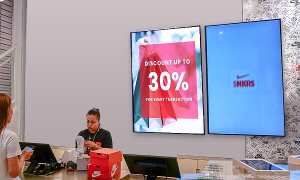 An employee bills Nike shoes at a pos billing counter while the shopper looks at an ad playing on large digital signs