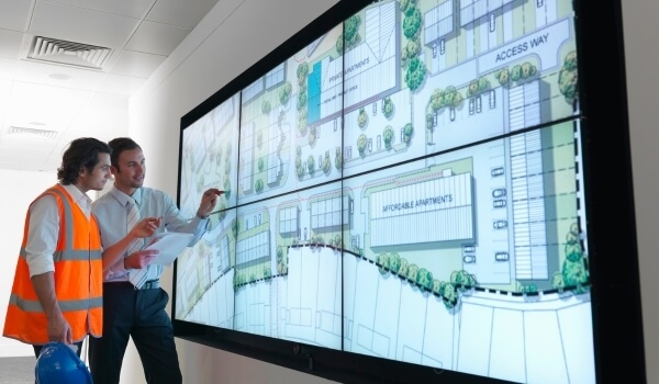 Two men point to a large real estate map shown on screen as they discuss construction plans