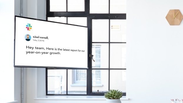 A digital screen installed inside an office shows the message from a team leader on Slack