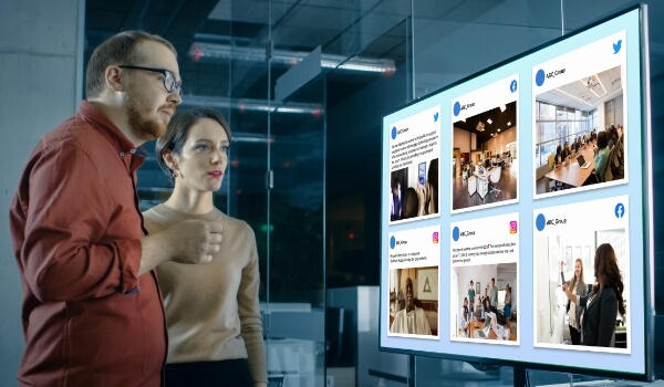 Employees inside an office break room stare at a TV display showing posts from Facebook, Twitter & Instagram