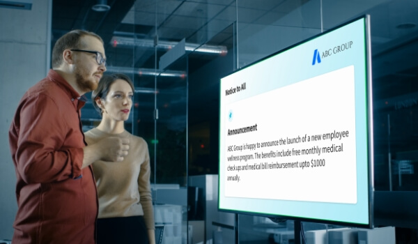 Two employees at an office read the notices displayed on a digital bulletin board