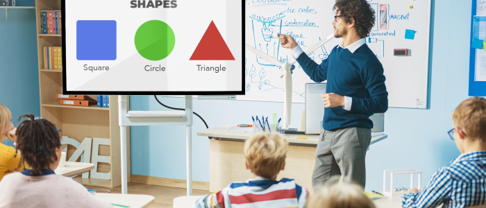 A kindergarten teacher shows bright colored shapes on a classroom digital signage as the students look at the screen.