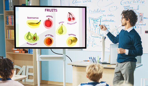 An elementary classroom digital signage shows bright & colorful images of fruits. A teacher uses the image content on screen to teach the students